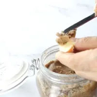 Almond butter in a glass jar. The almond butter is being spread onto a slice of apple.