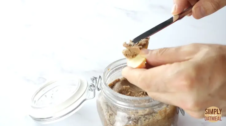 Almond butter in a glass jar. The almond butter is being spread onto a slice of apple.