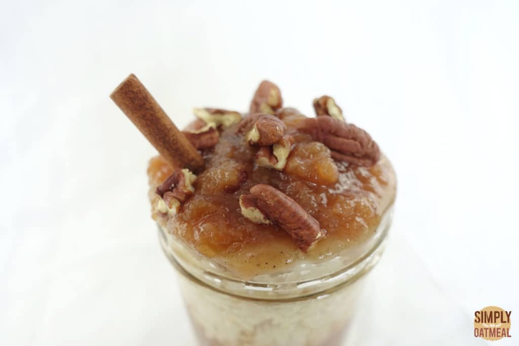 Apple cider overnight oats topped with applesauce, chopped pecans and a cinnamon stick in a glass bowl.
