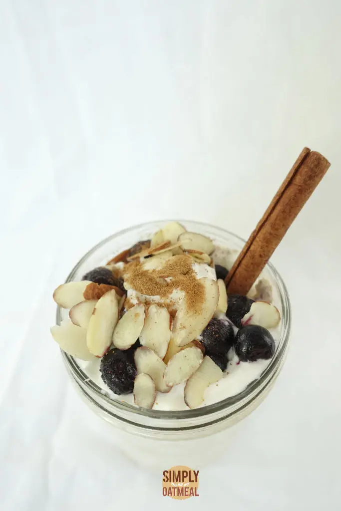 Top view of a mason jar filled with apple pie overnight oats. The oatmeal toppings include slivered almonds, fresh blueberries and a cinnamon stick.