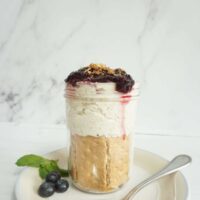 Mason jar filled with one serving of blueberry cheesecake overnight oats. The soaked oatmeal is layered with graham crackers and blueberry jam on top.