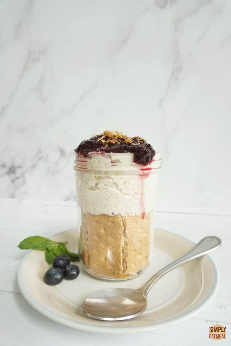 Mason jar filled with one serving of blueberry cheesecake overnight oats. The soaked oatmeal is layered with graham crackers and blueberry jam on top.