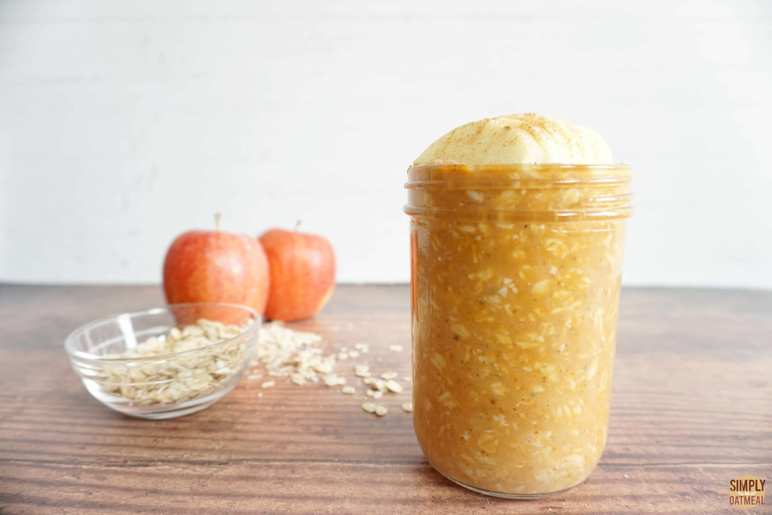 Pumpkin apple overnight oats in a glass container. 2 whole apples and small bowl of raw oats in the background.