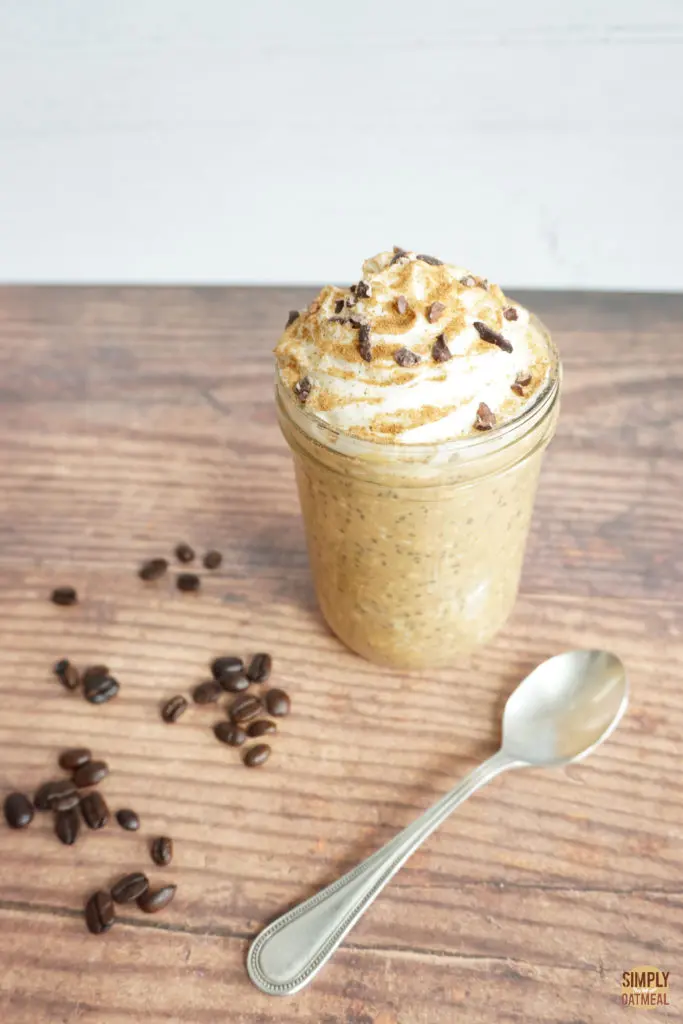 Glass jar with a serving of pumpkin spice latte overnight oats. A spoon and roasted coffee beans are on the wood table.