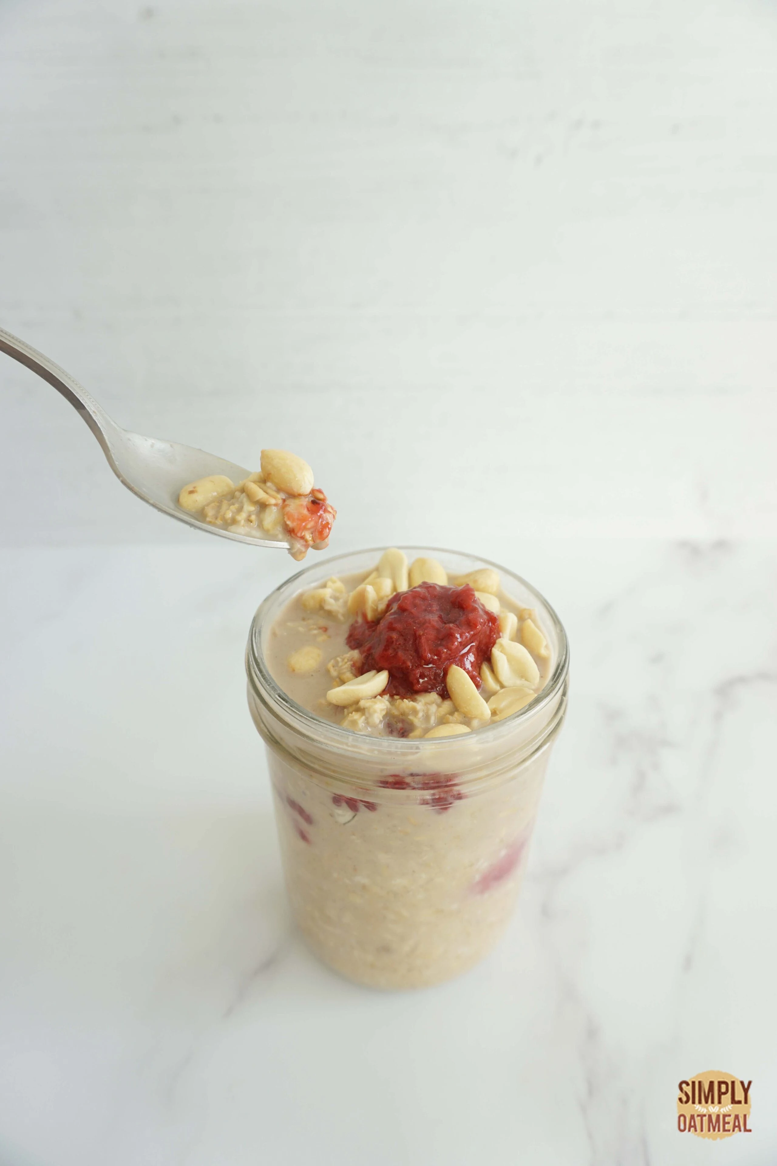 Raspberry peanut butter overnight oats served in a mason jar with a spoon scooping a bite. Toppings include crushed peanuts, homemade peanut butter and fresh raspberries.